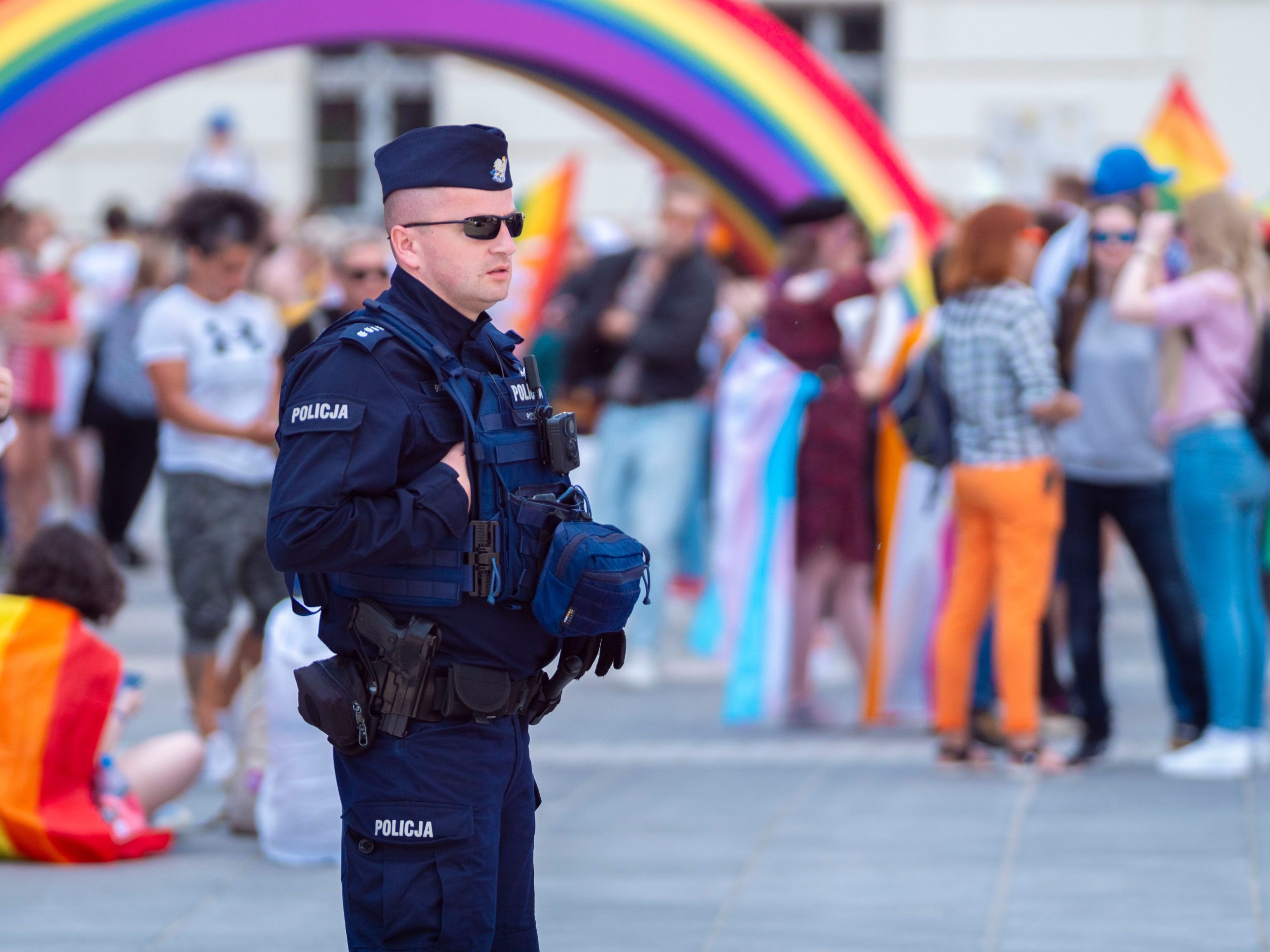 Police officer at a Pride parade in Wrocław, Poland, ensuring safety with people and rainbow flags in the background.